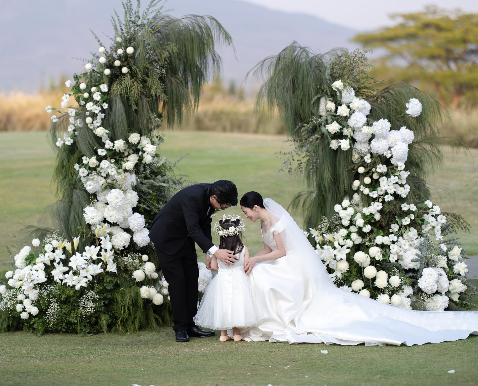 A couple kneeling to hug a little flower girl at their outdoor ceremony in Thailand, between tall white floral arches